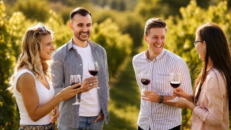 Four friends in a vineyard share wine and laugh together outdoors, each holding a glass of red wine.