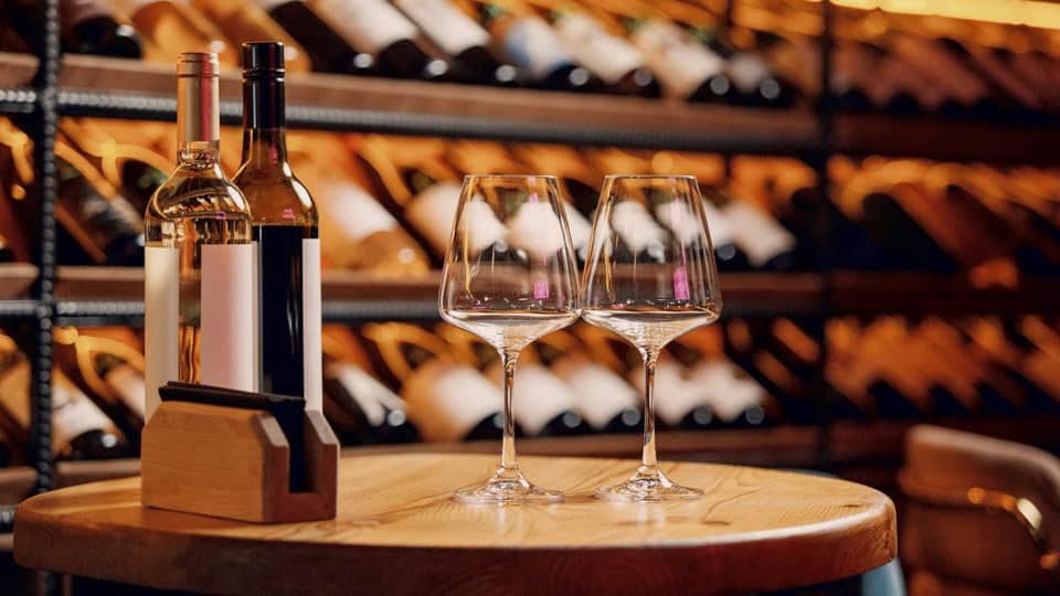 Two wine glasses and three bottles on a round wooden table in a warm wine cellar backdrop of shelves filled with bottles.