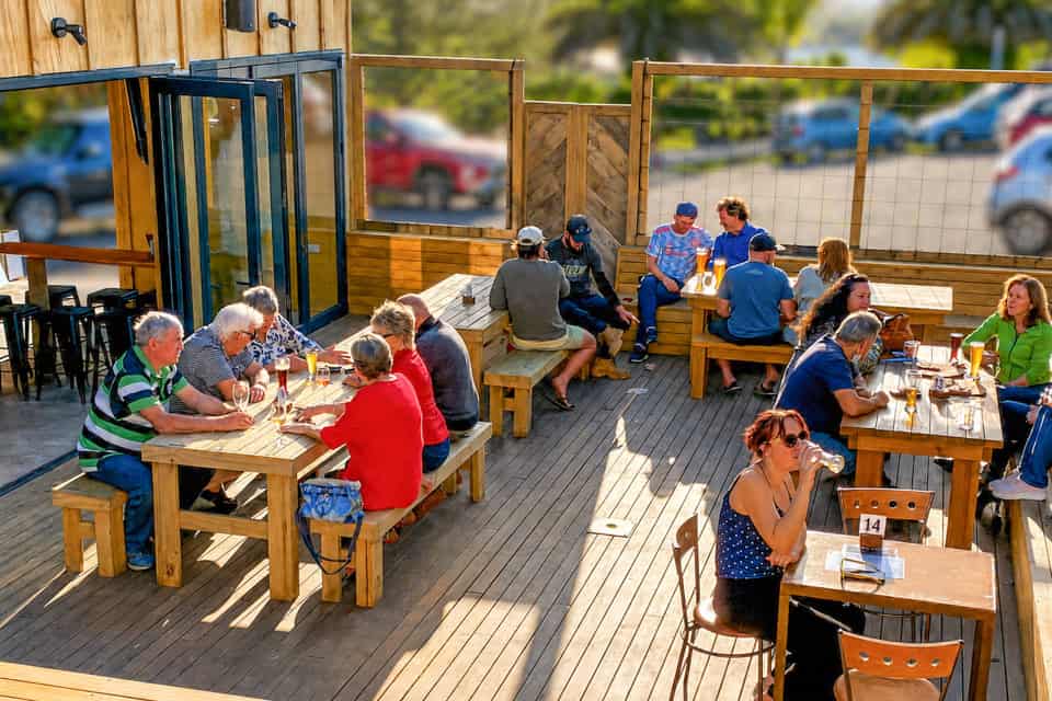 Groups of adults seated at long wooden picnic tables on a sunny outdoor deck, eating and drinking together nearby a doorway.