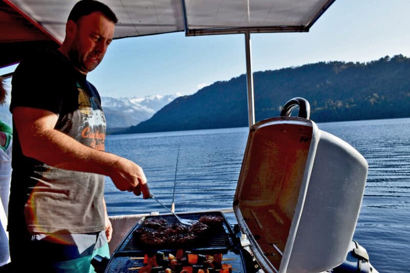 Man grilling burgers on a barbecue aboard a boat on a calm lake with mountains in the background.