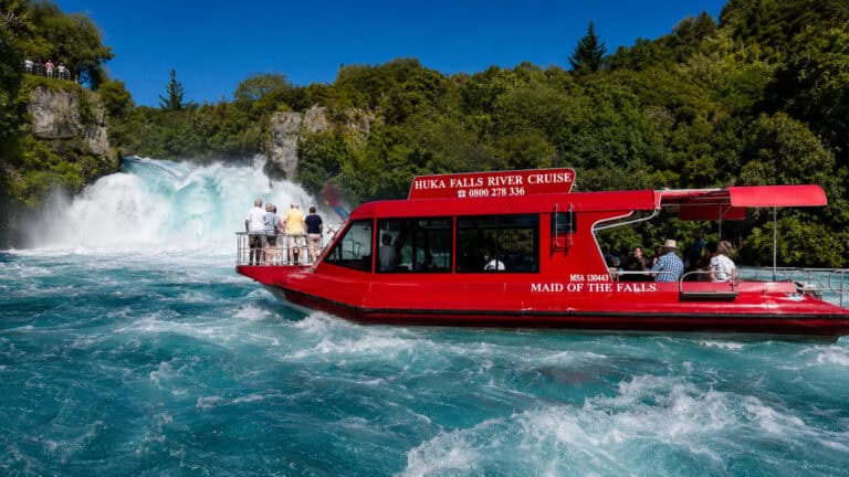 Red passenger boat a Huka Falls on the waikato river Taupo with onlookers on the riverbank cliffs and lush green trees in the background.