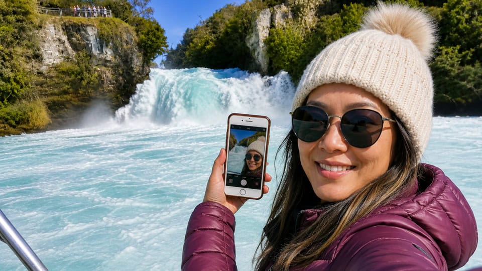 Woman in a burgundy jacket and cream knit hat smiles while taking a selfie with a smartphone, a Huka Falls waterfall behind her at Lake Taupo