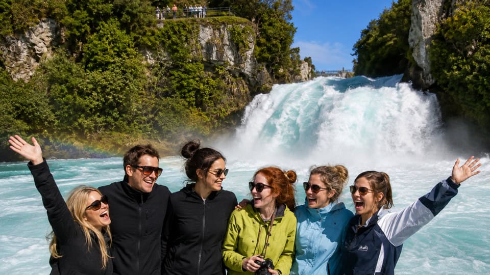 Six friends posing with arms raised in front of a cascading waterfall and turquoise river behind them.