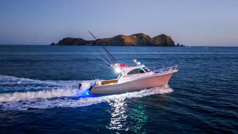 White motorboat speeding across blue ocean, island in distance, with wake and deck lights reflecting on the water.