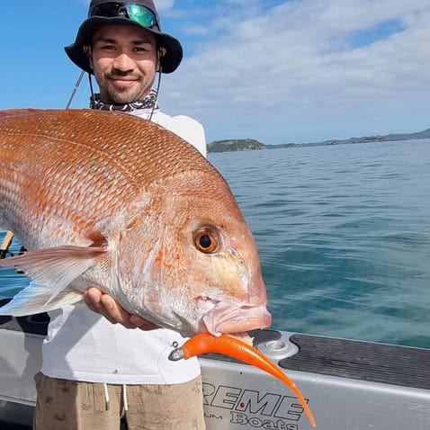 Man on a boat holds a large red snapper with an orange lure in its mouth, blue water and distant islands in the background.