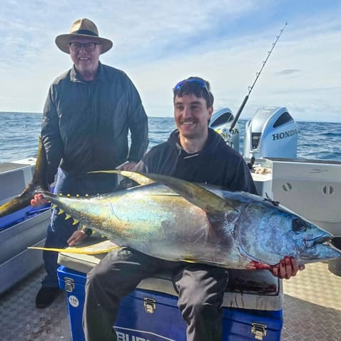 Two men on a boat pose with a huge yellowfin tuna perched on a cooler, sea in the background.