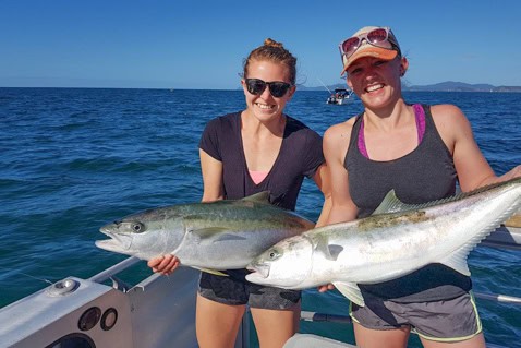 Two women on a boat posing with two large fish they just caught, the ocean and blue sky behind them.