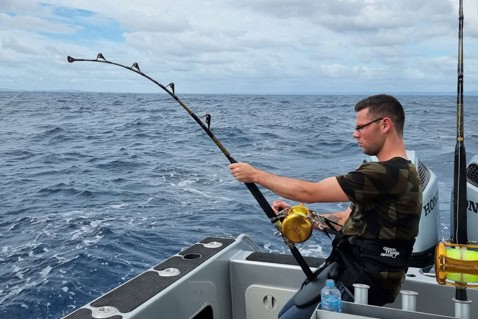 Man in camouflage shirt seated on a boat, fishing rod raised over the open ocean.