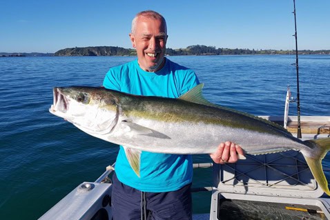 Smiling man on a boat holding a large yellowtail fish against a calm blue sea.