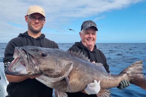 Two men on a boat proudly hold a large fish across the frame against an ocean backdrop.