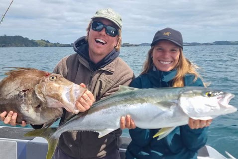 Two people on a boat holding a large fish they just caught, smiling at the camera, with blue water and distant shoreline in the background.