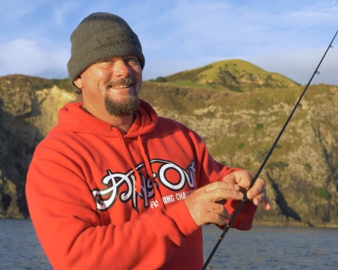 Smiling man in an orange hoodie and green beanie fishing along a rocky coast.