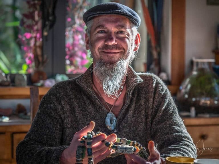 Rakiura-Jade-stewrt-island-15 | Must Do New Zealand | Must Do New Zealand Smiling man with gray beard and flat cap, holding colorful beaded bracelets indoors in a cozy greenstone carving workshop on Stewart Island