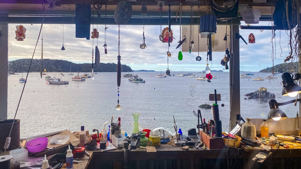 Cluttered workshop bench with scattered tools and hanging trinkets, viewed through a window at a harbor with boats from the workspace interior.