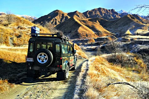 Off-road SUV with a spare tire on the back drives a dusty dirt road through sunlit desert hills and mountains.