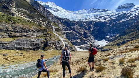 Three hikers with backpacks stand on a rocky meadow facing a snow-capped glacier in a rugged mountain landscape.