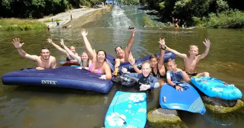 Group of friends smiling and waving from a large blue Intex inflatable raft on a river, with boards nearby and a small waterfall in the background.