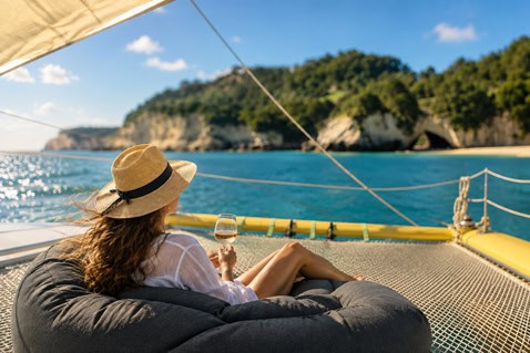 Woman in a sun hat relaxing on a bean bag on a boat, sipping wine with blue sea and rocky cliffs in the background.