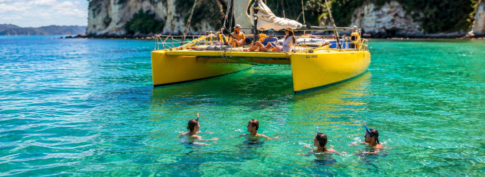 People swim near a bright yellow catamaran in turquoise water by a rocky shoreline. The boat sits close to the swimmers, with others on board enjoying the day.