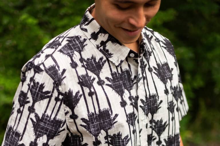 Man wearing a white shirt with a black palm tree pattern, outdoors with greenery in the background