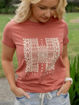 Woman standing outdoors wearing a pink T-shirt with a bold white geometric graphic on the chest, hands in her pockets.