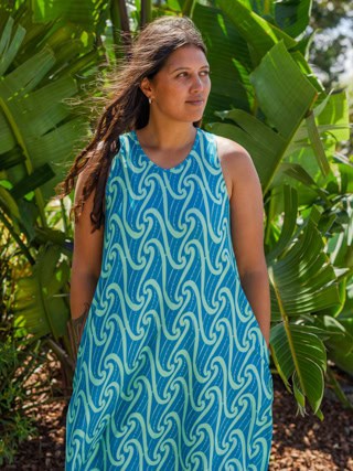 Woman wearing a blue sleeveless dress with a wavy pattern, standing among tropical green plants outdoors, looking to the side.