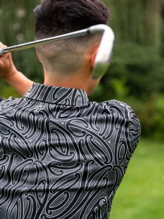 Person viewed from behind, mid-swing with a golf club on a green golf course, wearing a patterned shirt.