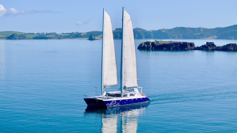 Two-masted sailing yacht with white sails gliding on calm blue water, with rocky coastline in the distance.