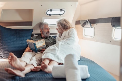 Older couple in robes lounging on a bed inside a camper, reading a book together.