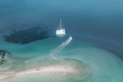 Aerial view of a white sailboat anchored near a sandy beach with clear turquoise water and a small group of people on shore.