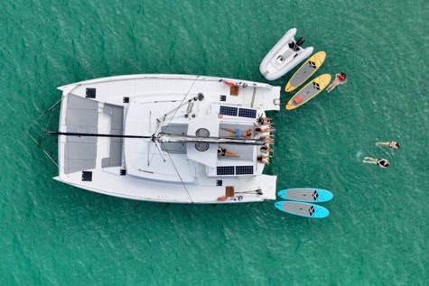 Aerial view of a white catamaran anchored in turquoise water with people on deck and multiple paddleboards nearby.