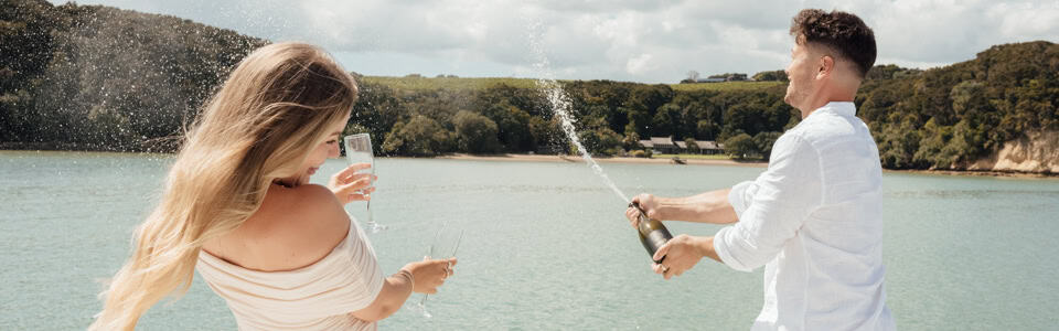 Couple celebrating by a lake: woman with long blonde hair toasting with a glass while a man in a white shirt sprays champagne toward her.