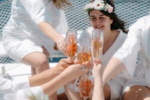 Group of friends toasting with champagne glasses outdoors, wearing white outfits and a flower crown—celebration on a sunny day.