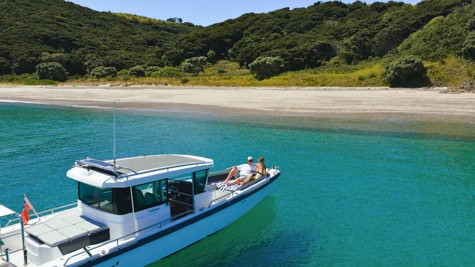 A white motorboat with two people relaxing on a turquoise bay, near a sandy beach and green trees in the background.