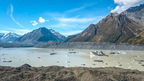 Snowy mountains surround a pale blue glacial lake with floating ice chunks under a bright blue sky.