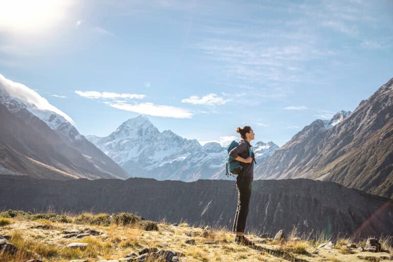 Hiker with a teal backpack stands on a grassy rocky foreground, with snow?capped mountains and a clear blue sky behind.