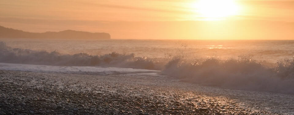 Sunset over a pebbled beach with waves crashing and a distant silhouetted coastline on the horizon, warm orange glow across the water and shore.