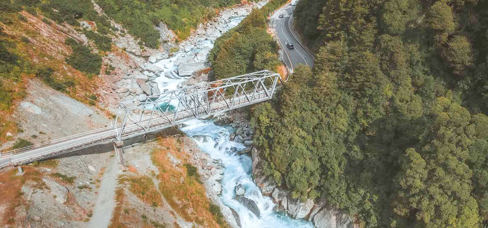 Gate of Haast. Aerial view of a metal truss bridge spanning a rushing river in a forested gorge, with a winding road and cars on the right side.