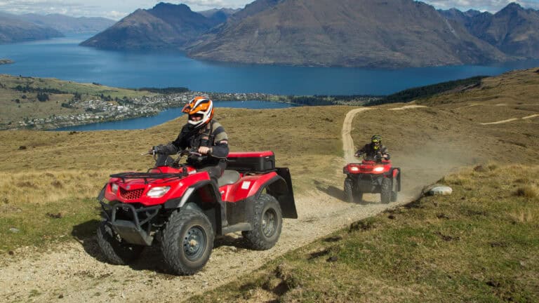 Two riders on red ATVs travel a dusty hillside trail with a blue lake and towering mountains in the background.