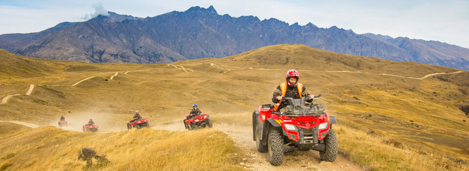 Rider in an orange vest on a red ATV leading a dusty trail across golden hills with mountains in the distance.