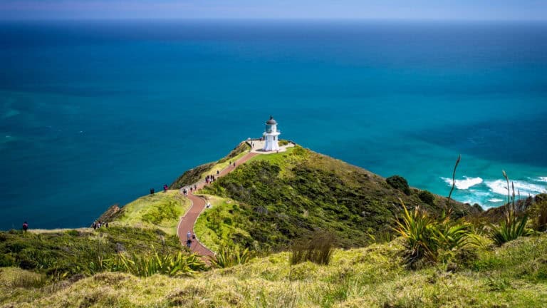 White lighthouse on a grassy headland with a winding path and visitors, overlooking a blue ocean.