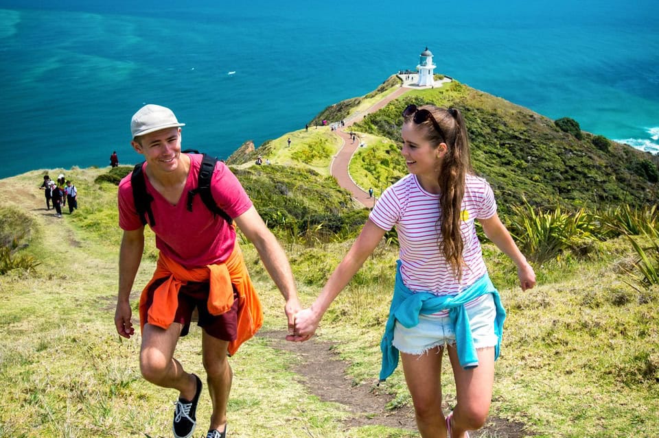 Couple hiking up a grassy coastal trail holding hands toward a white lighthouse and blue ocean.