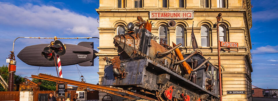 Exterior of Steampunk HQ with a giant rusty mechanical sculpture on a crane-like installation in front, blue sky above. Oamaru