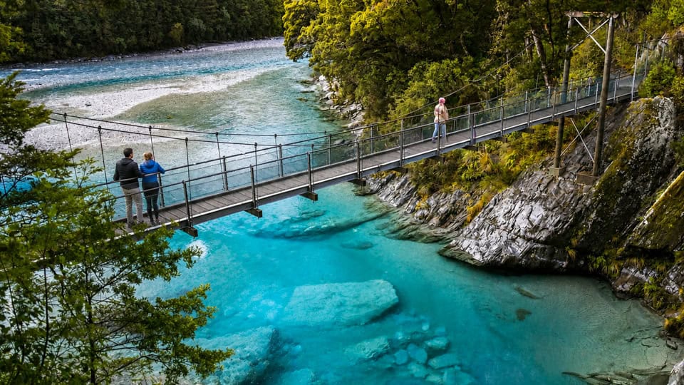 Suspension bridge crossing a turquoise river with rocky banks, three people walking along and enjoying the view.