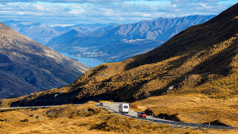 Winding mountain road with a white coach and a red car, golden hills in the foreground and blue alpine mountains in the distance.