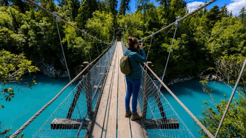 A person in a teal jacket with a backpack stands on a wooden suspension bridge and photographs the turquoise river below amid lush green trees.