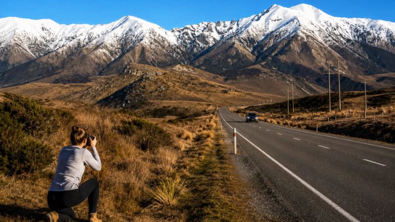 Photographer kneeling in brush taking a photo of snow-capped mountains across a rural highway at golden hour