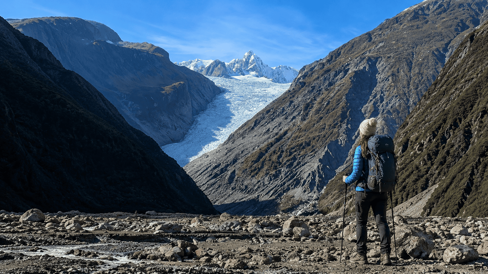 Hiker in a blue jacket and beige beanie stands with trekking poles, gazing at a glacier between tall mountain walls.