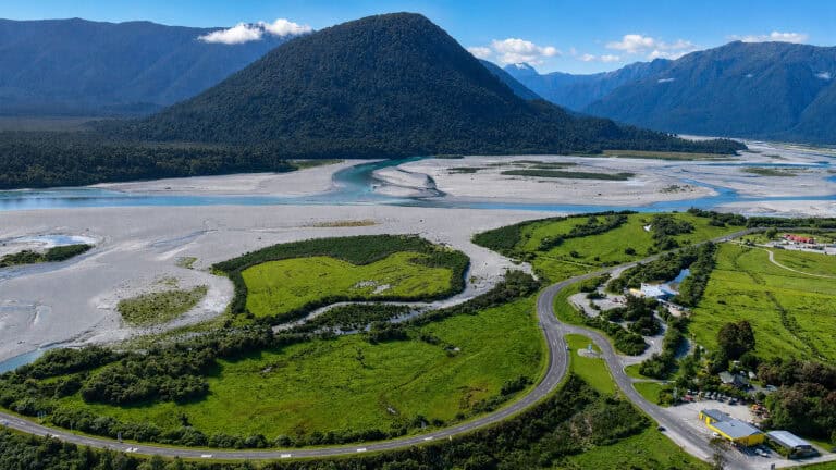 Haast-Township-22 | Must Do New Zealand | Must Do New Zealand Haast Aerial view of a winding highway through green fields beside a braided river delta with forested mountains in the background.