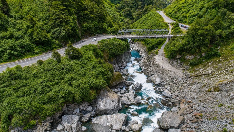 Aerial view of a rocky river gorge with turquoise water, dense green trees, and a road with a metal bridge crossing above.